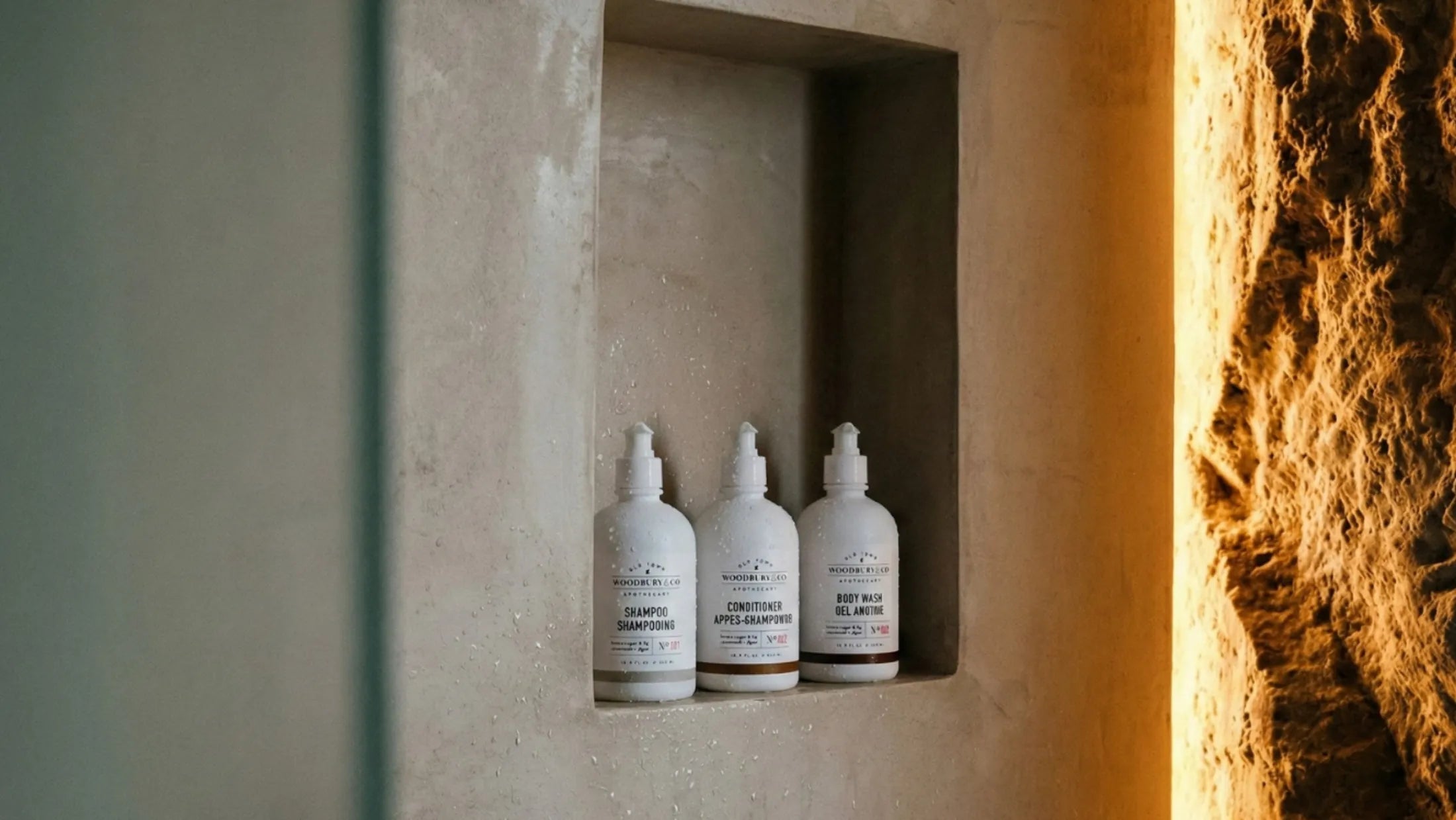 Woodbury & Co shampoo, conditioner and body wash bottles arranged in a sunlit stone bathroom alcove
