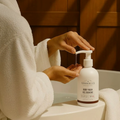 Person in a white robe using a pump bottle of Woodberry & Co. body wash in a bathroom.