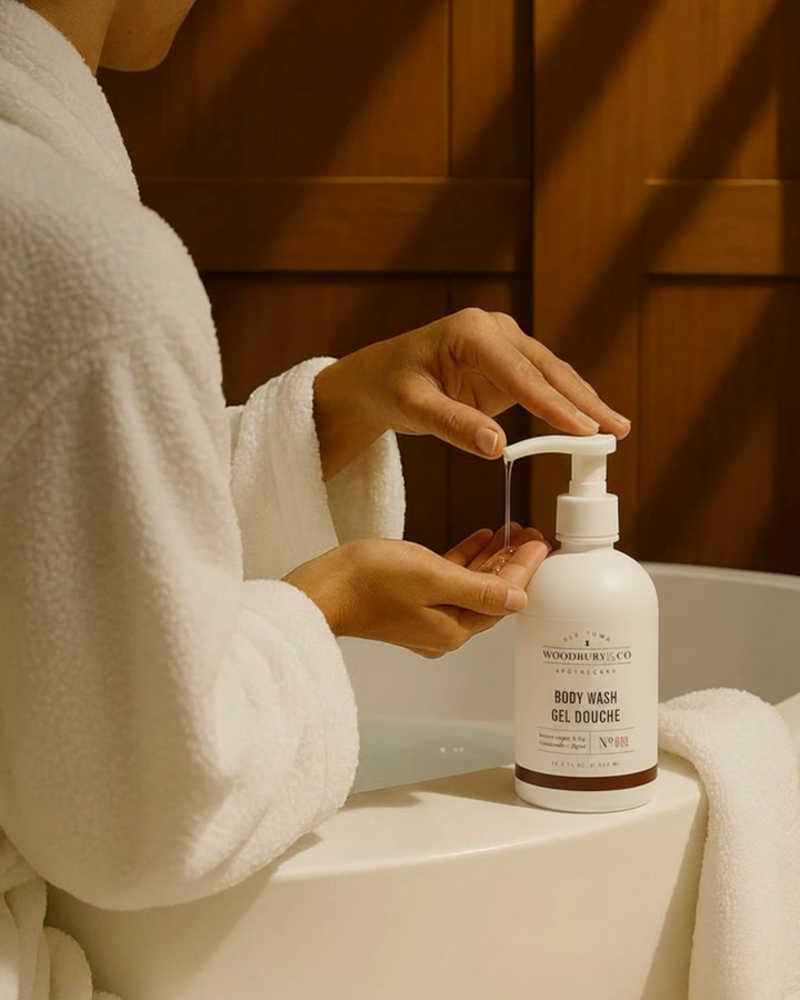Person in a white robe using a pump bottle of Woodberry & Co. body wash in a bathroom.