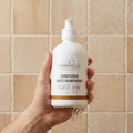 Hand holding a bottle of Woodbury & Co. conditioner against a tiled shower wall.