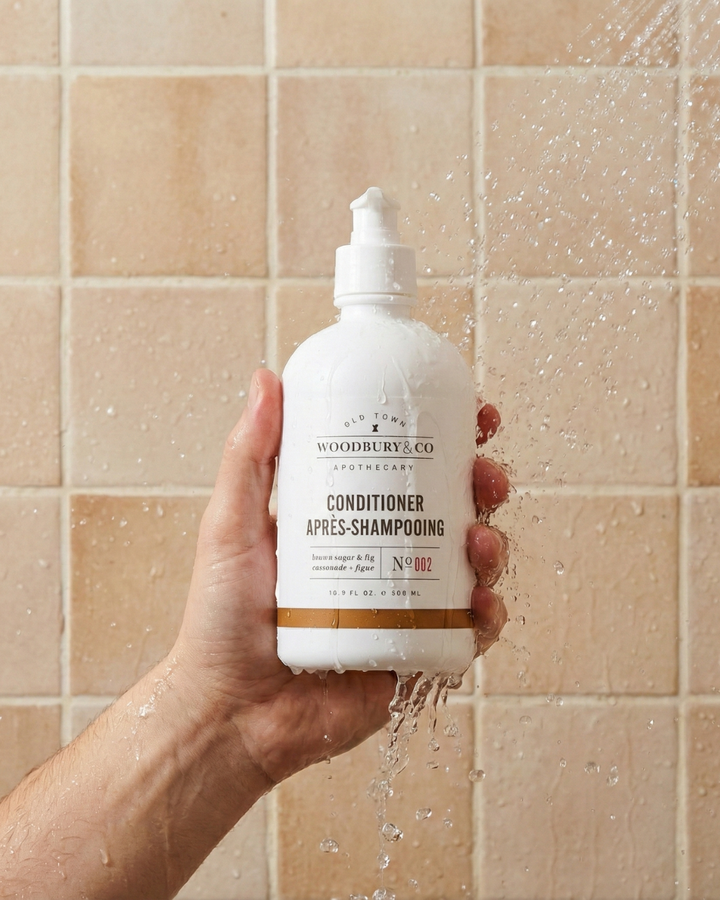 Hand holding a bottle of Woodbury & Co. conditioner against a tiled shower wall.