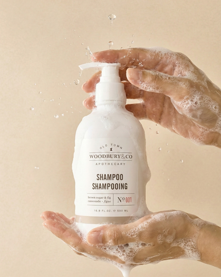 Bottle of Woodbury & Co. shampoo held by hands with soap suds on a beige background